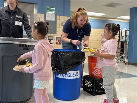 two girls standing in front of recycle can with a woman helping to recycle their items