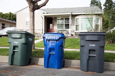 A green trash bin, blue trash bin and black trash bin outside of a white house