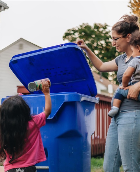 A little girl throwing something into a blue recycle bin with a woman standing by holding a baby.